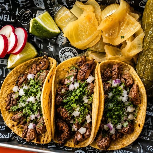 Three tacos with meat, cilantro, and onions, served with radishes, limes, and pickled vegetables.