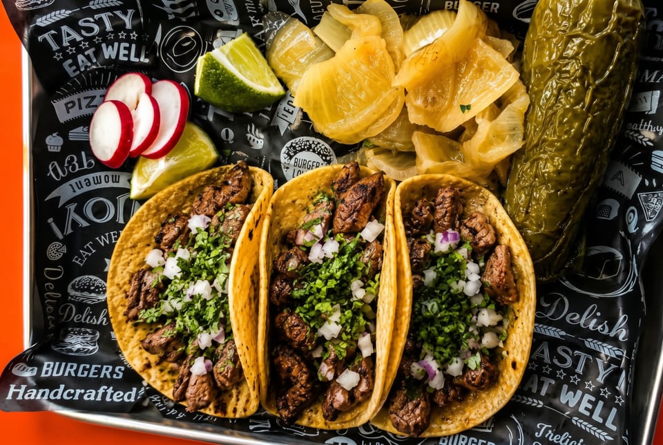 Three tacos with meat, cilantro, and onions, served with radishes, limes, and pickled vegetables.