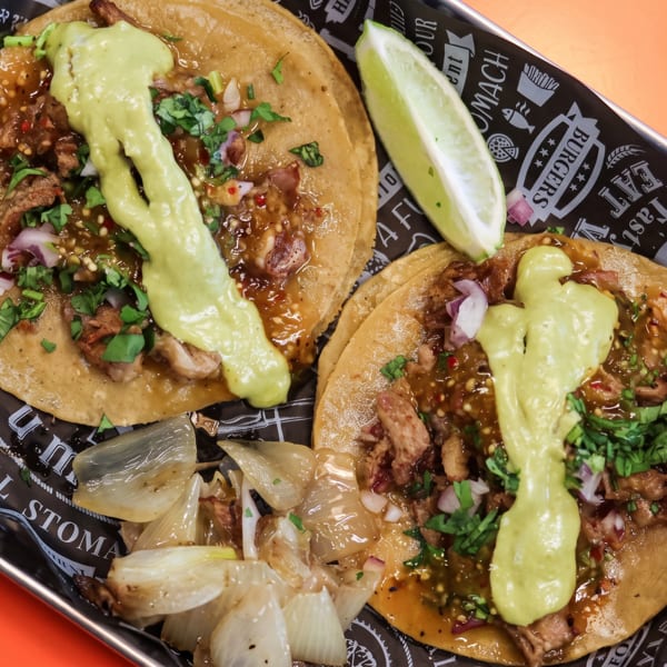 Two street tacos with meat, salsa, avocado crema, cilantro, and onions, with a lime wedge.