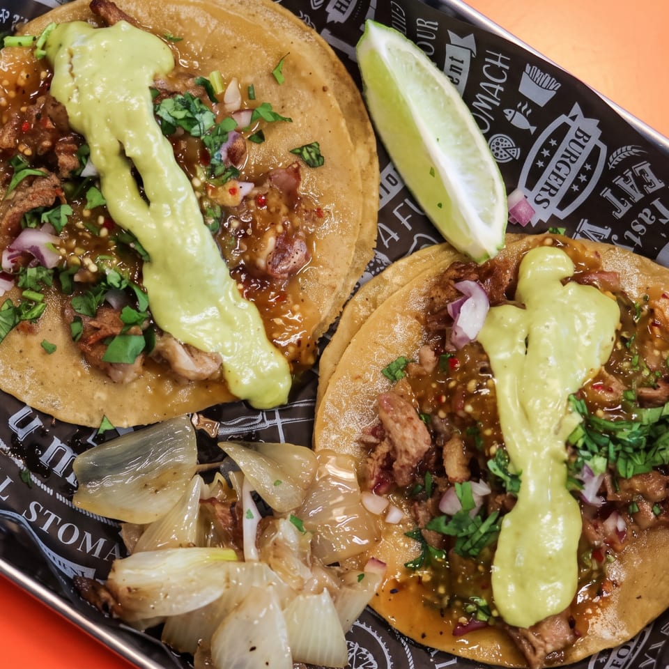 Two street tacos with meat, salsa, avocado crema, cilantro, and onions, with a lime wedge.