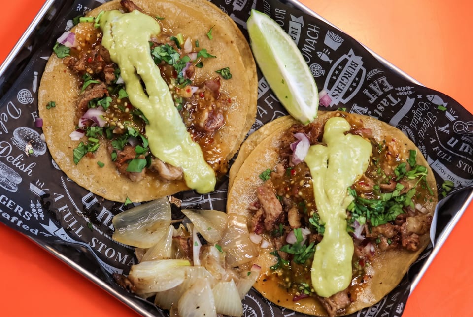Two street tacos with meat, salsa, avocado crema, cilantro, and onions, with a lime wedge.