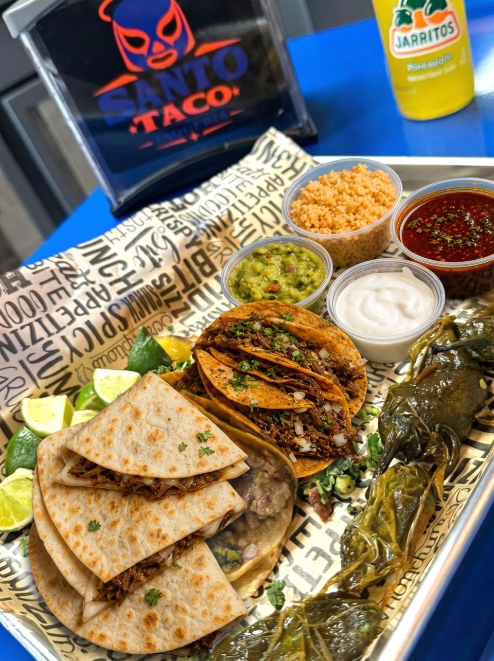 Santo Taco birria tacos, quesadillas, and sides on a tray with a Jarritos pineapple soda.