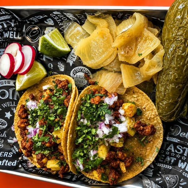 Two tacos with meat, pineapple, cilantro, and onions, served with radishes, limes, pickled onions, and a large pickle.