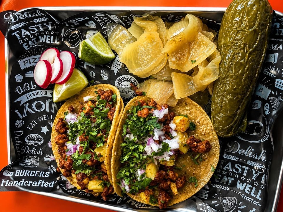 Two tacos with meat, pineapple, cilantro, and onions, served with radishes, limes, pickled onions, and a large pickle.