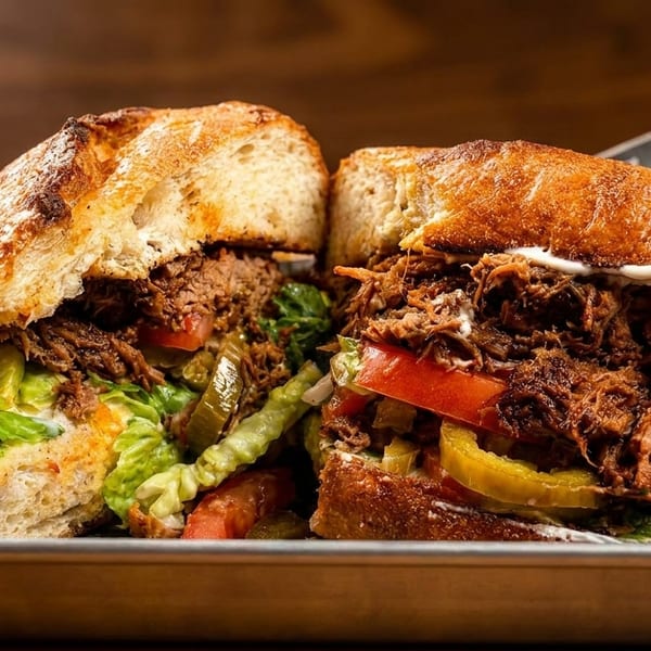 Two halves of a large, crusty bread sandwich filled with shredded meat, lettuce, tomatoes, and pickles.