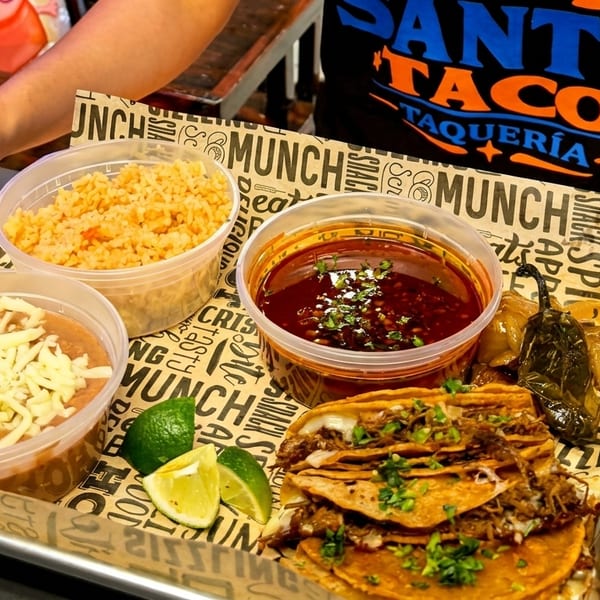 A tray of Santo Taco birria tacos, rice, beans, consommé, and limes on a branded paper liner.