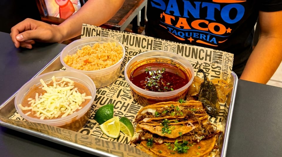 A tray of Santo Taco birria tacos, rice, beans, consommé, and limes on a branded paper liner.