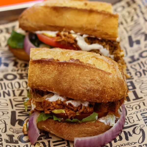 Two pulled pork sandwiches with lettuce, tomato, onion, and sauce on a branded paper liner.