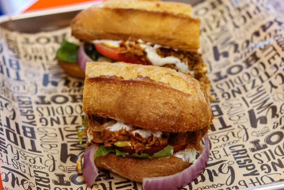 Two pulled pork sandwiches with lettuce, tomato, onion, and sauce on a branded paper liner.