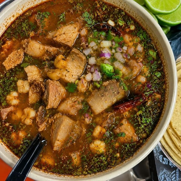 Hearty bowl of pozole with pork, hominy, cilantro, and diced onions, served with lime wedges.