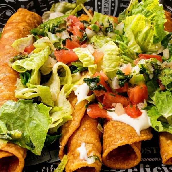 Crispy taquitos topped with fresh lettuce, diced tomatoes, and sour cream on patterned paper.