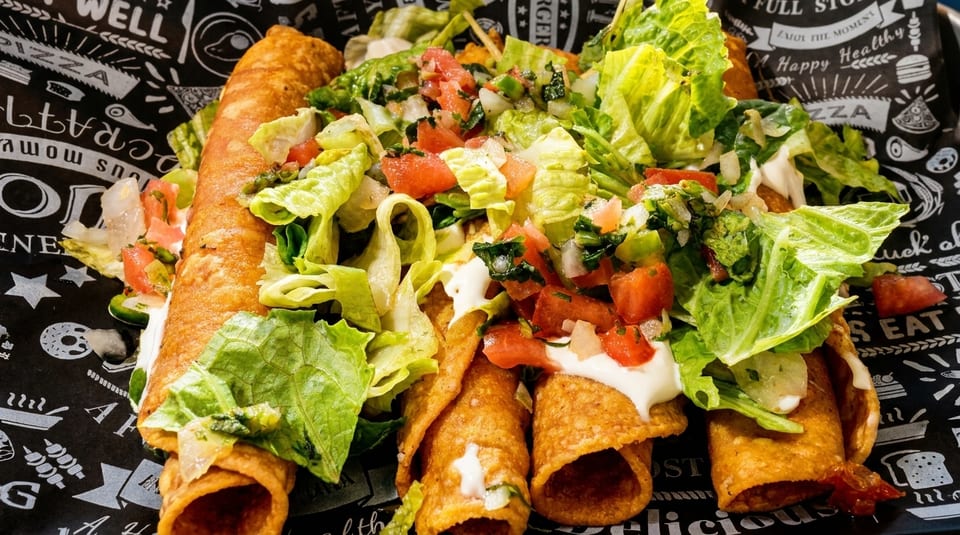 Crispy taquitos topped with fresh lettuce, diced tomatoes, and sour cream on patterned paper.