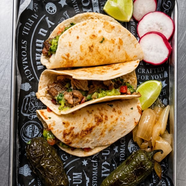 Three tacos with guacamole and meat, served with radishes, limes, and peppers on a metal tray.