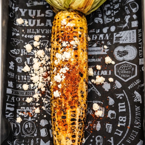 Grilled corn on the cob with cotija cheese and chili powder on black patterned paper.