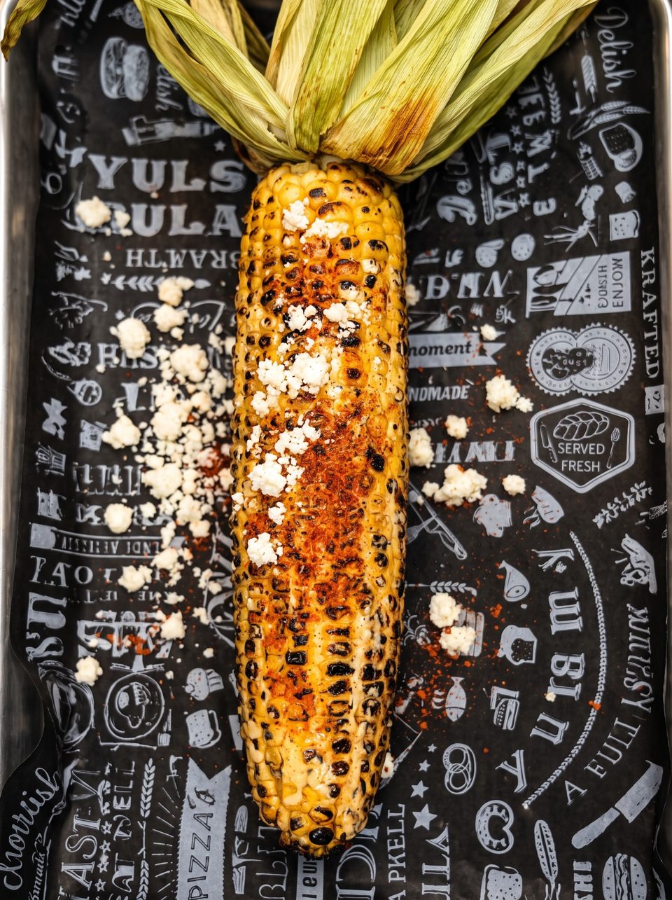 Grilled corn on the cob with cotija cheese and chili powder on black patterned paper.