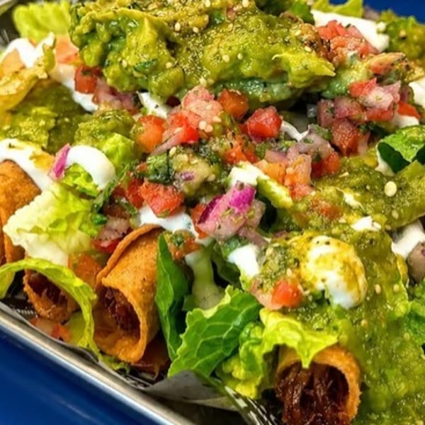 Crispy taquitos topped with guacamole, salsa, sour cream, and fresh lettuce in a metal tray.
