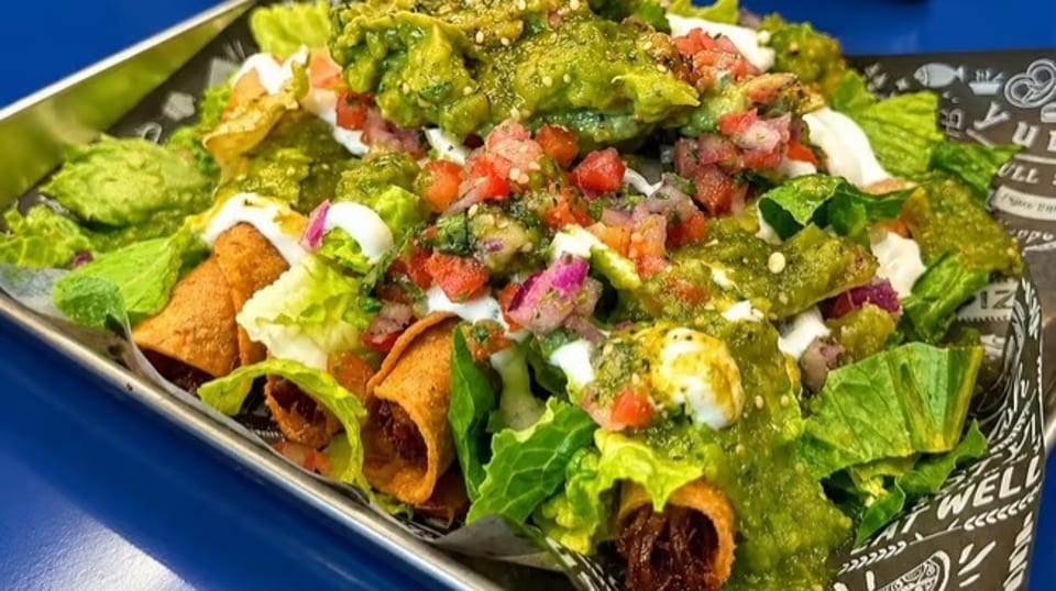 Crispy taquitos topped with guacamole, salsa, sour cream, and fresh lettuce in a metal tray.